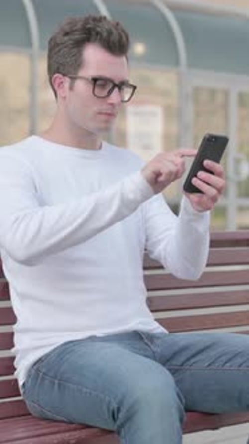 Young Man Using Smartphone while Sitting Outdoor on Bench, Vertical Video