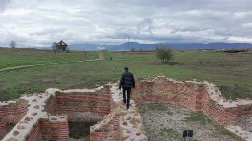 Aerial view of a man walking through ancient city ruins in a large green field on a cloudy day