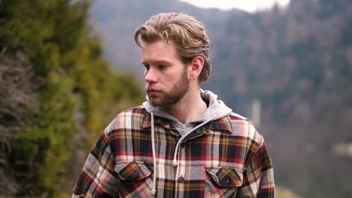 Ein junger blonder Mann in einem karierten Hoodie geht an einem Fluss entlang, wobei sich sein zerzaustes Haar und seine blauen Augen verfangen