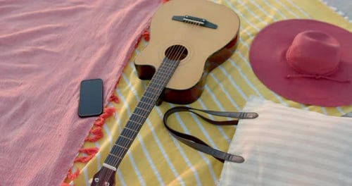 A guitar lies next to a hat and phone on a beach blanket