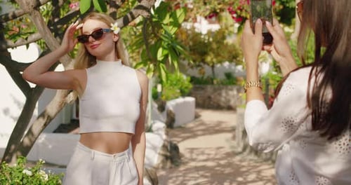 Woman posing with sunglasses and a flower in her hair, standing in a garden, being photographed.