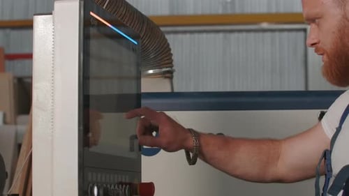 Man with beard in work uniform at factory. Man adjusts control panel of industrial machines