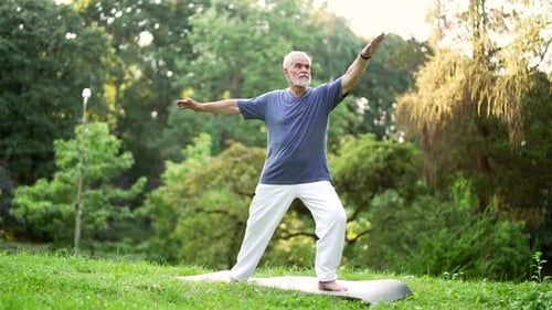 Senior active gray haired bearded man does gymnastics while standing on a mat in an urban city park.