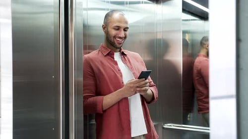 Smiling businessman is using mobile phone while standing in the elevator in modern office building.