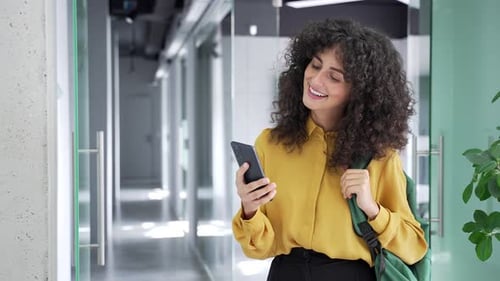 Smiling female student is using mobile phone standing at university indoors. Happy curly brunette