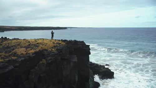 Wide shot of man taking photos of waves from cliffside in Iceland