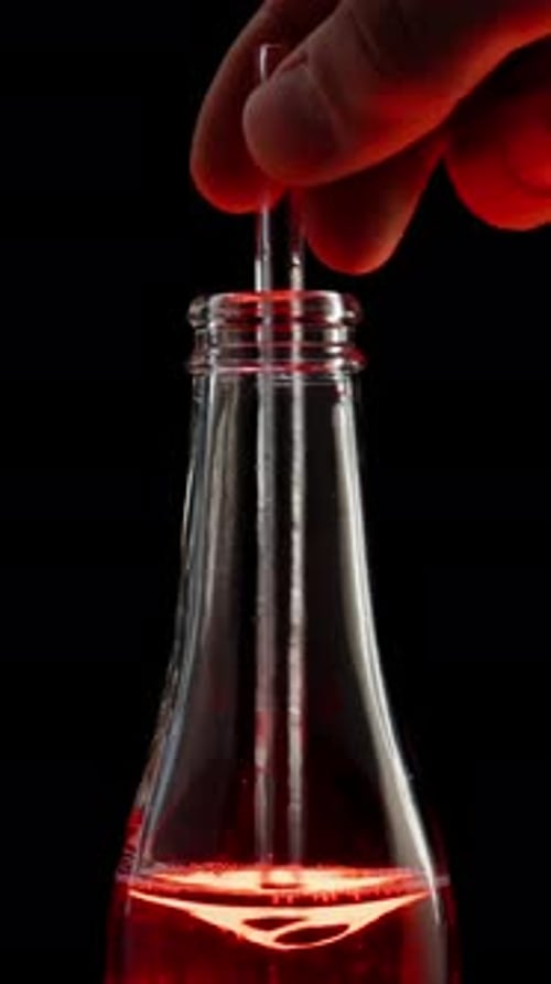 Vertical video. Red soda in a glass bottle is stirred with a transparent straw, creating a vortex.