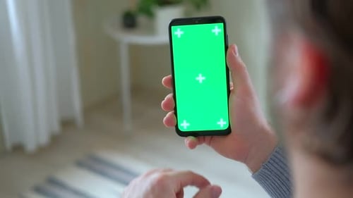 Young Man Sitting at Home Holding Smartphone Green Mockup Screen in Hand Male Person Using Chroma