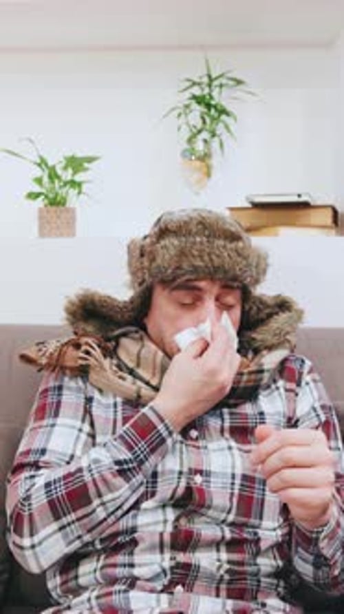 An Elderly Grayhaired Man in a Warm Earflap Hat Sneezes and Coughs Into a Napkin While Having the