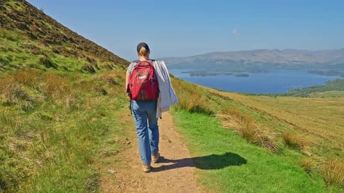 Following a hiker walking on a mountainside trail surrounded by grassy fields and majestic views