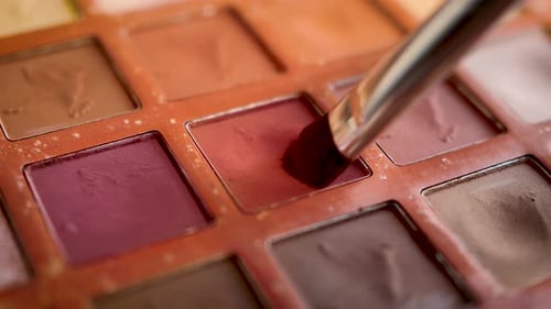 A close-up of a cosmetic, multi-colored, eyeshadow makeup palette as a brush applicator is expertly