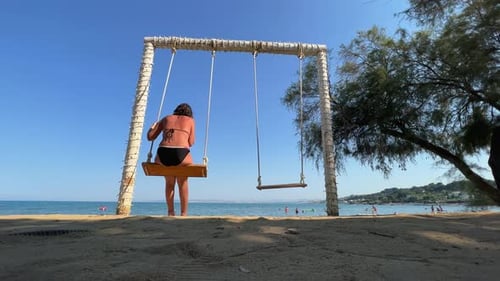 Low angle back view of adult woman on summer holiday enjoy swinging on rope swing looking at calm se