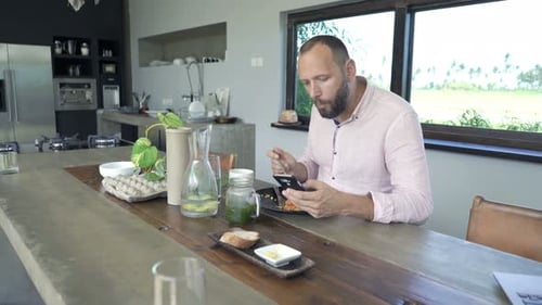 Man Eating and Using Phone at Kitchen Table