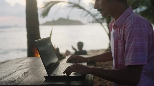 Mann in Freizeitkleidung arbeitet am Laptop im Strandcafé mit Meerblick bei Sonnenuntergang Freelancer-Typen