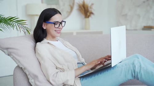 Young Woman Relaxes on Sofa Working on Laptop