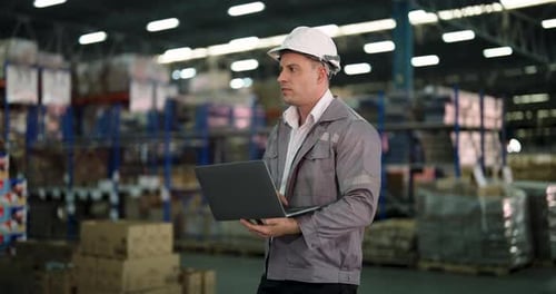 Männlicher Ingenieur steht in einem Lagerhaus, umgeben von Pappkartons und Regalen, hält Laptop und CH
