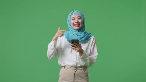 Asian Muslim Woman Smiling And Pointing To Smartphone In Her Hand In The Green Screen Background