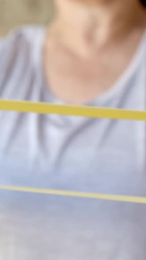 Vertical video: Placing yellow band on wrist, woman in grey tee pulling left arm at home for rehab