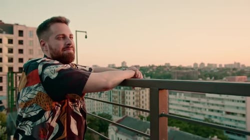 ein junger bärtiger Mann steht auf einer Dachterrasse und schaut in die Stadt und denkt über die Zukunft nach