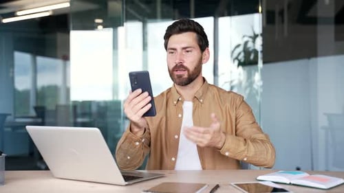 Glücklicher Geschäftsmann, der mit einem Handy in einem Videoanruf spricht, während er bei der Arbeit am Schreibtisch sitzt