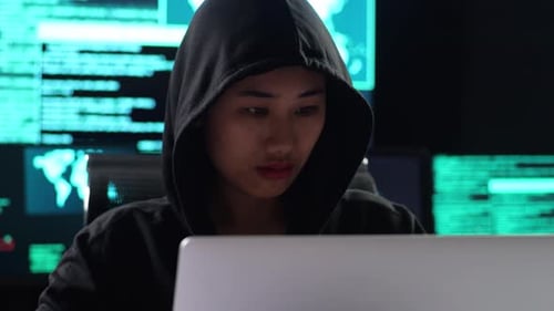 Close Up Of Asian Female Hacker In A Hood Works On A Laptop With Multiple Monitors In A Dark Office