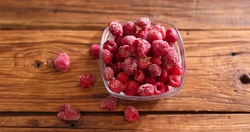 Ripe frozen raspberries and bowl on wooden table, camera moving forward