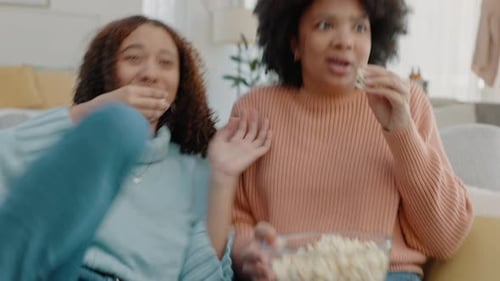 Women friends, watching tv and scary movie, film and media in lounge at home while eating popcorn