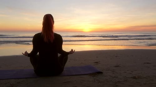woman meditating while performing lotus flower sitting looking at sea during sunrise