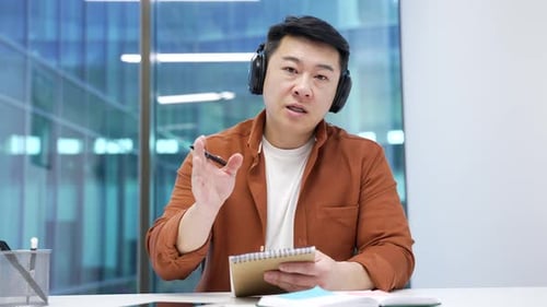 Webcam view. Asian businessman in wireless headphones watching video call conference notes