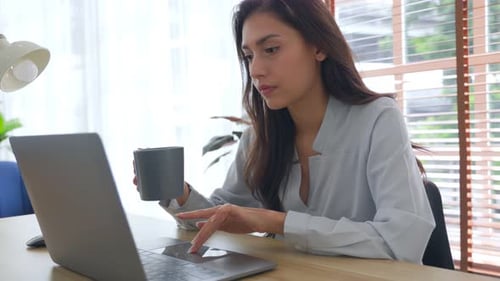 Frau arbeitet am Laptop und trinkt Kaffee im Heimbüro.