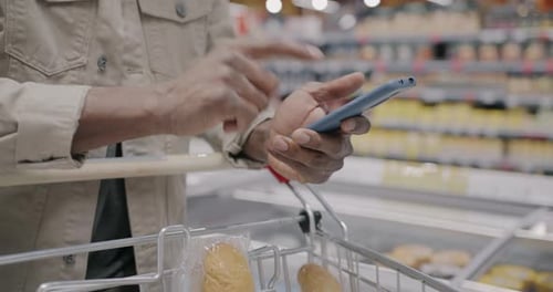 Closeup of Male Hand Using Smartphone and Checking Products in Shopping Trolley While Man Buying