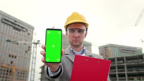 Engineer Presents Smartphone with Green Screen at Construction Site