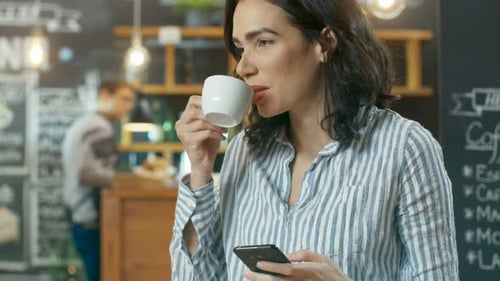 Beautiful Woman Uses Smartphone while Sitting in Cafe. She Drinks Beverage from the Cup. Background
