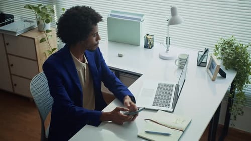 African American Businessman Checking Smartphone in front of Laptop at Office