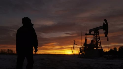 Worker Silhouette in Hood Comes to Pumpjack Against Sunset