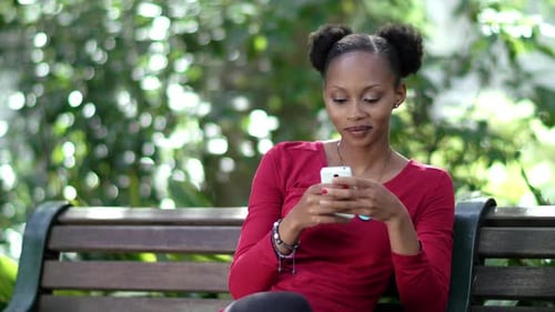 4k Video footage of a young woman sitting on a bench in a park sending text messages on her phone