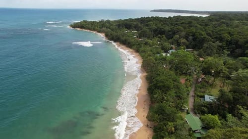 Flight over a paradisiacal beach with turquoise water bordered by a jungle with some huts.