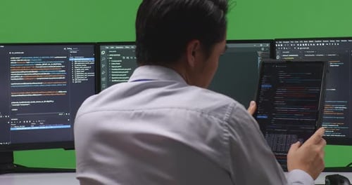 Developer Working With Tablet While Write Code With Multiple Computer Screens In Green Screen