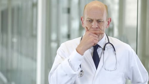 A pensive male doctor is standing in a hospital corridor thinking and pondering. The senior man is w