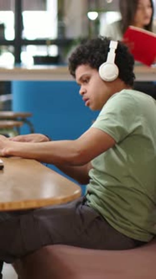 Vertical video: Putting on white headphones, student in green tee studying at lounge with laptop
