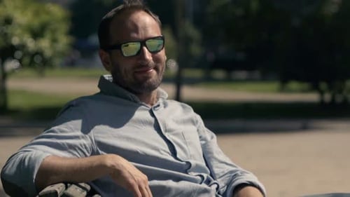 Smiling young man relaxes on a bench in a sunny city park on a summer day
