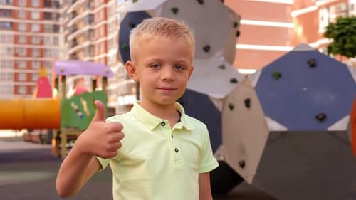 A Little Boy Stands on the Playground Looks at the Camera and Shows a Thumbs Up