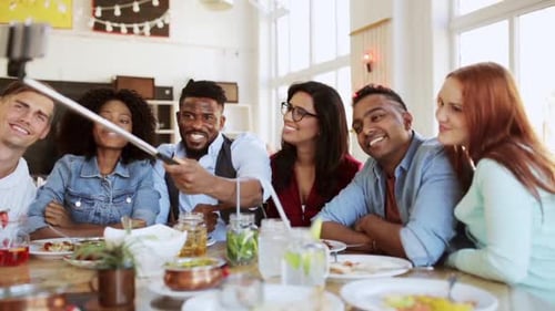 Happy multiethnic friends taking smartphone selfies at dinner in a restaurant