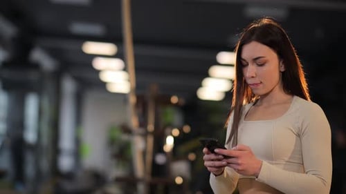 Young Attractive Woman with a Beautiful Figure Looks at a Phone in the Gym