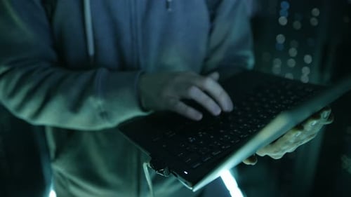 Hooded Hacker Typing on Laptop in Data Center