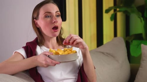 Young Woman is Relaxing on the Couch Watching TV and Eating Chips
