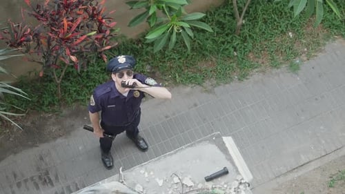Police officer is standing in a building yard, looking up holding radio to his mouth.
