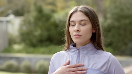 Woman placing hand on chest inhaling to calm opening eyes smiling in garden with athletic jacket