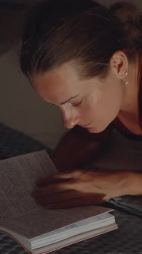 At night, the young woman lies on the edge of the bed and looks at the notebook, top view Vertical,