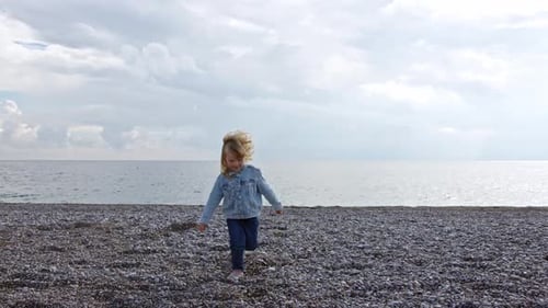 Small Threeyearold Child Run Away From Cold Sea on Camera Along Stone Beach in Denim Jacket and